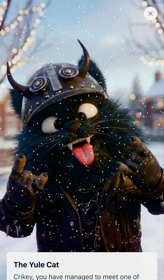 The Yule Cat, a scruffy black cat in a horned helmet and leather jacket, sticking its tongue out in the snow.